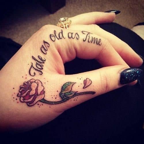 Elegant hand tattoo featuring a delicate rose with a falling petal on the finger and 'Tale as old as Time' scripted across the hand.