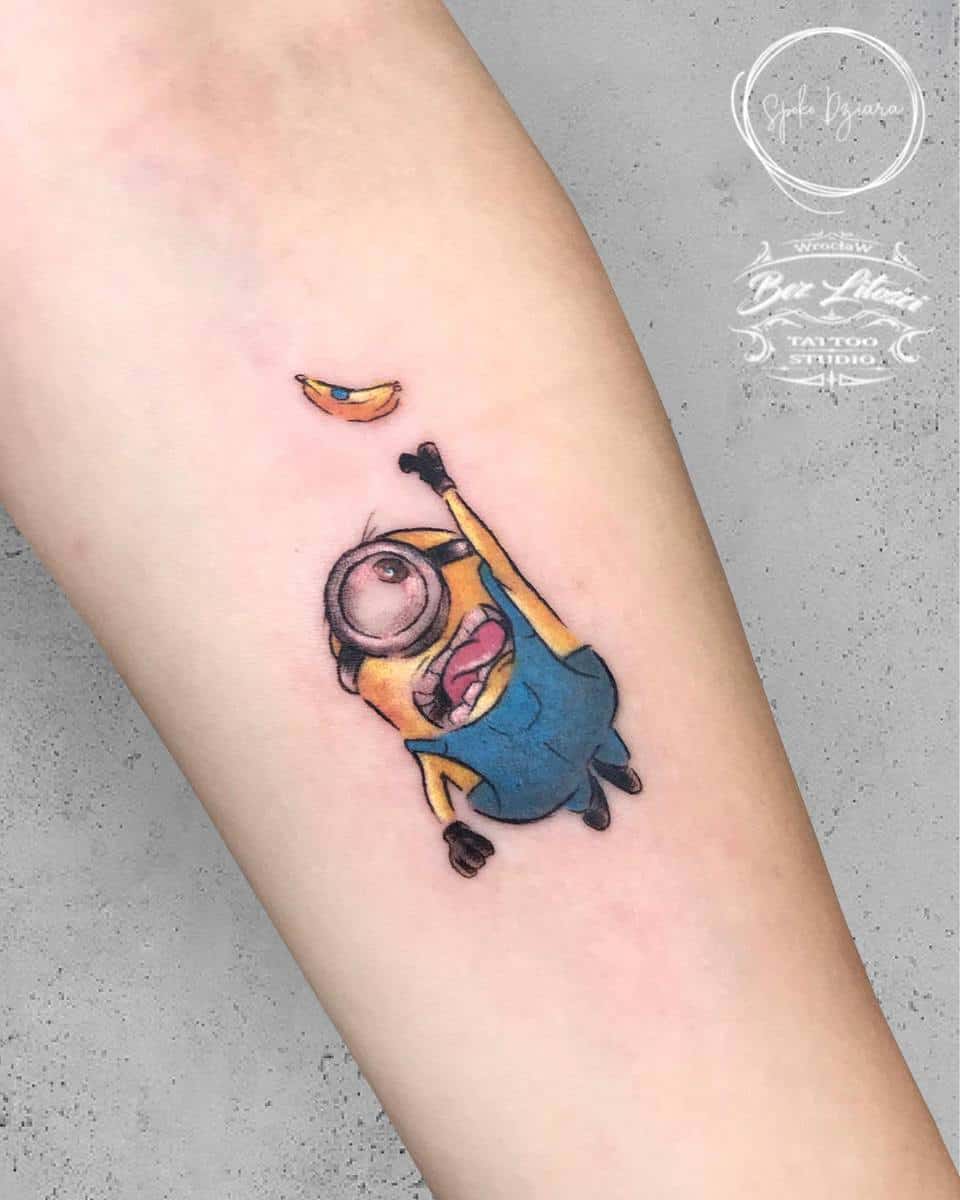 A cute tattoo of a Minion reaching for a small, floating banana on a forearm