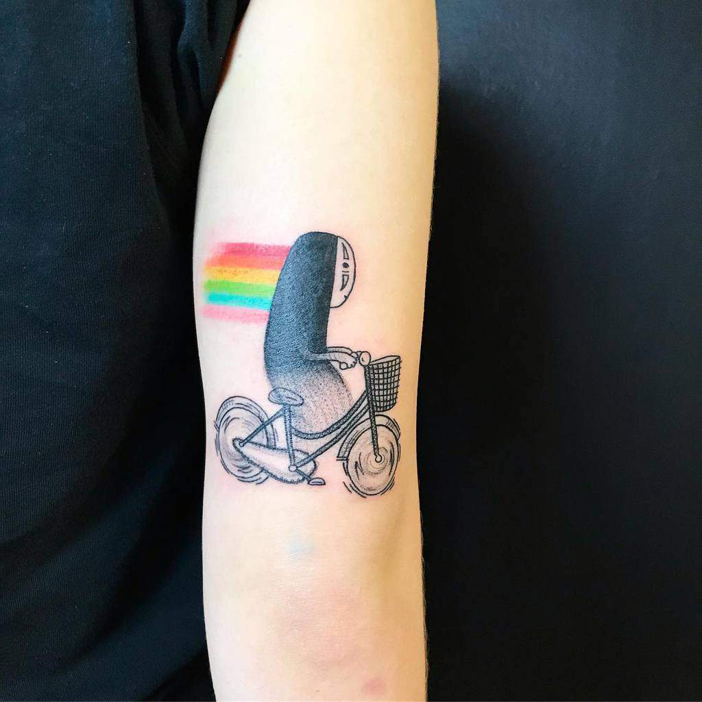 Playful tattoo of No-Face from Spirited Away riding a bicycle, with a rainbow trail behind, blending blackwork with colorful accents