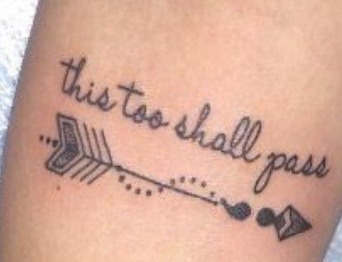 A "This Too Shall Pass" tattoo in cursive with a decorative arrow design, on a person's arm.