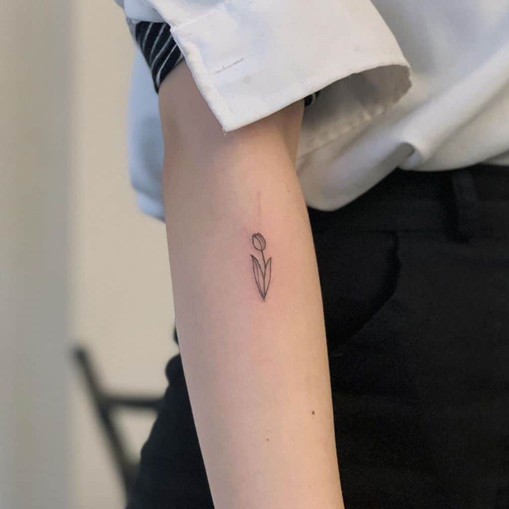 A minimalist tulip tattoo on the forearm, featuring a simple black-line design with a small flower and geometric leaves.
