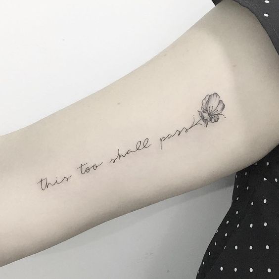 A "This Too Shall Pass" tattoo in cursive with a delicate flower design at the end, on a person's forearm.