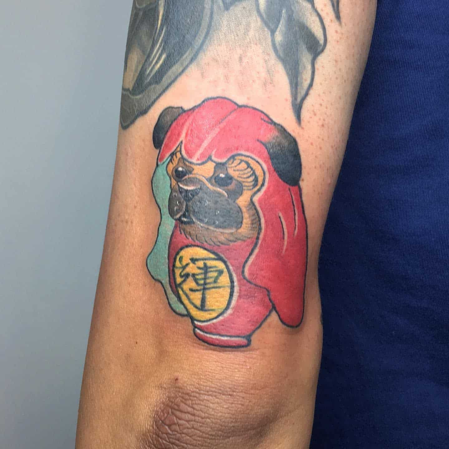 Tattoo of a pug in red attire resembling a Daruma doll with blue background on the side