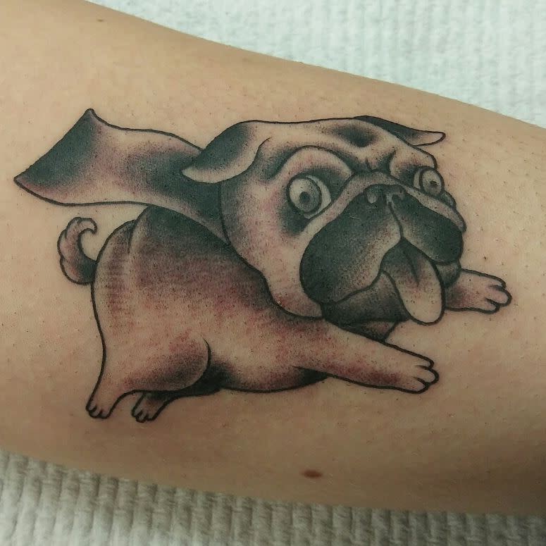 Tattoo of a cartoon-style pug with exaggerated eyes and tongue, with a cape