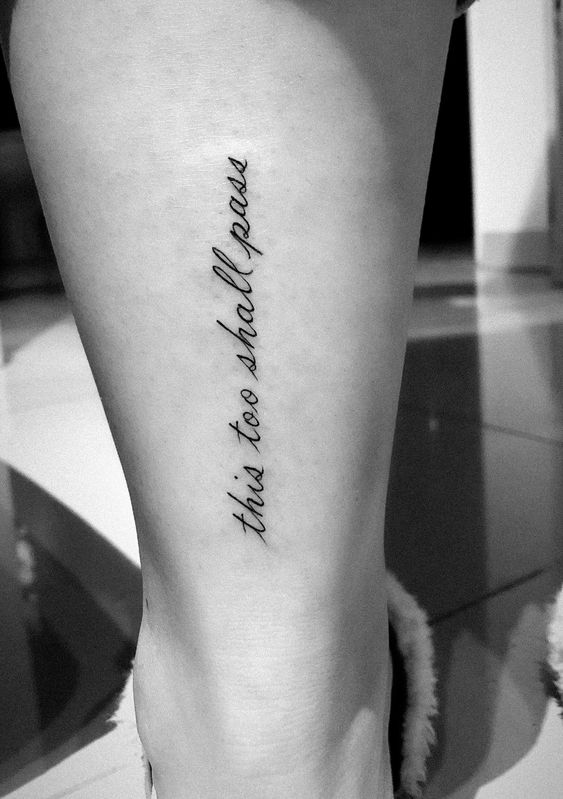 "this too shall pass" tattoo in cursive on the lower leg, in black and white.