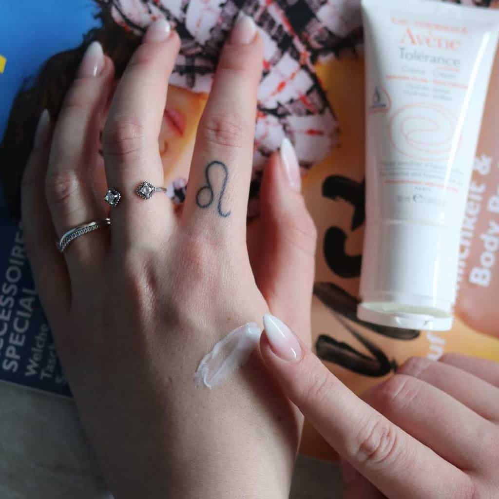 Minimalist black ink Leo symbol tattoo on the middle finger, paired with elegant rings.