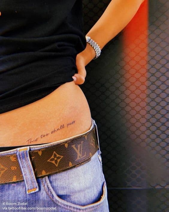 A "This Too Shall Pass" tattoo on a person's side, near the waist, with a Louis Vuitton belt visible in the frame.