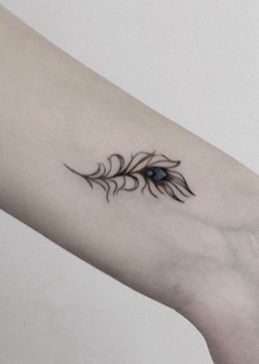 Small and delicate peacock feather tattoo on the wrist, featuring fine black linework with subtle blue shading in the eye pattern.