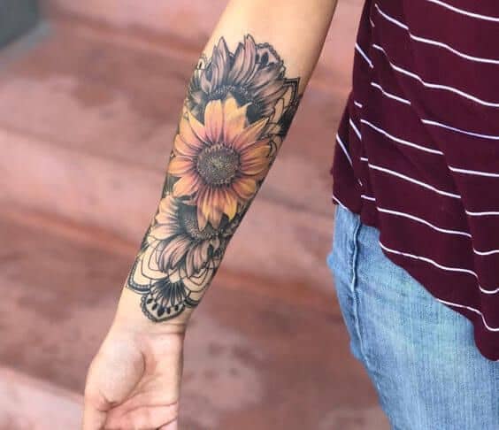 Forearm tattoo featuring a vibrant sunflower with warm orange petals, surrounded by black and grey floral and mandala-style details.