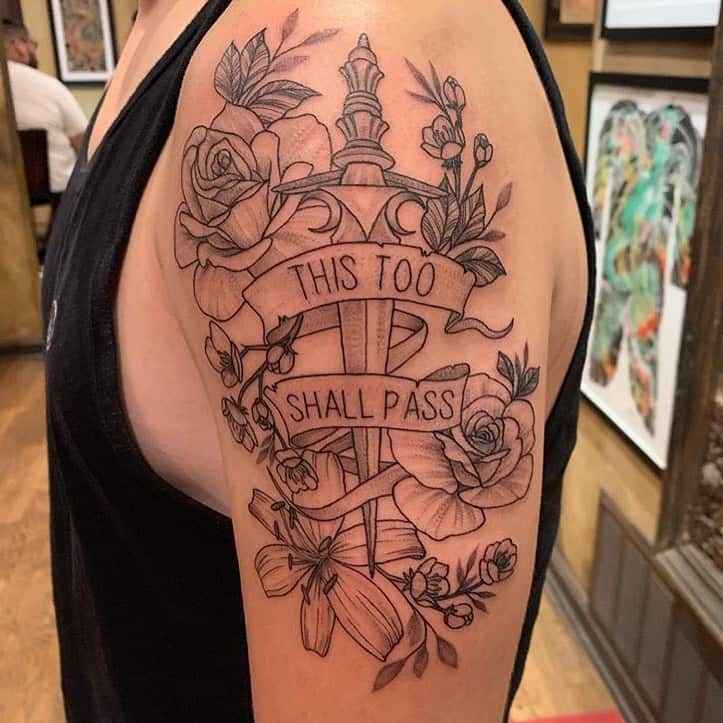 A bold "This Too Shall Pass" tattoo featuring a detailed sword intertwined with roses and banners, symbolizing resilience and strength. The placement on the upper arm adds to its striking presence.
