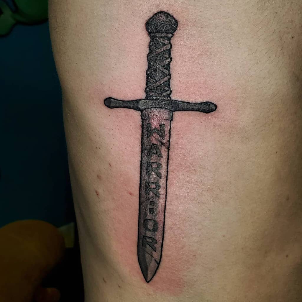 The tattoo features a sword with the word "WARRIOR" engraved on the blade, symbolizing strength and resilience. The design is bold and impactful with sharp, clean lines.