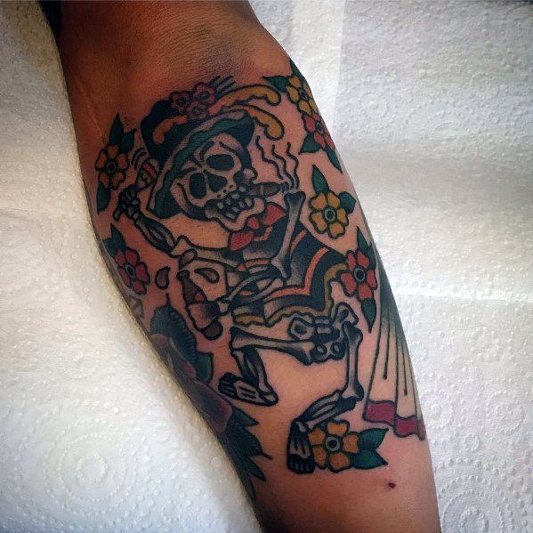 Colorful dancing skeleton tattoo in a festive outfit with a hat, surrounded by flowers, creating a lively and vibrant Día de los Muertos theme.