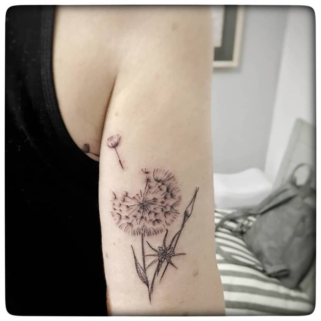 This tattoo depicts a detailed dandelion in soft black ink, capturing its delicate appearance along with some flower stems and a few dandelion seeds floating away. The design is minimalist yet expressive.