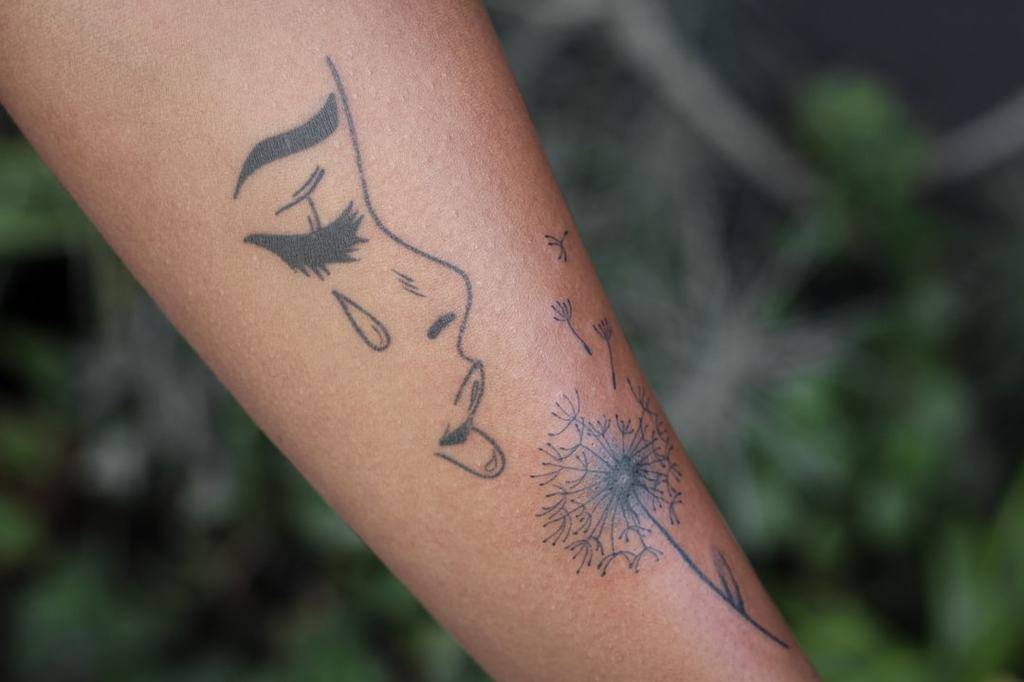 A delicate tattoo of a woman's face with a tear, beside a dandelion puff with seeds blowing away, on the forearm.
