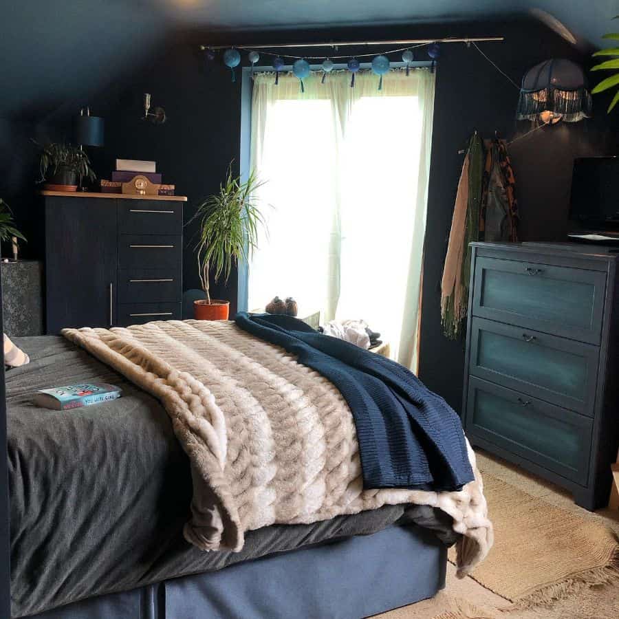 Cozy bedroom with dark blue walls, soft bedding, greenery, and vintage accents.