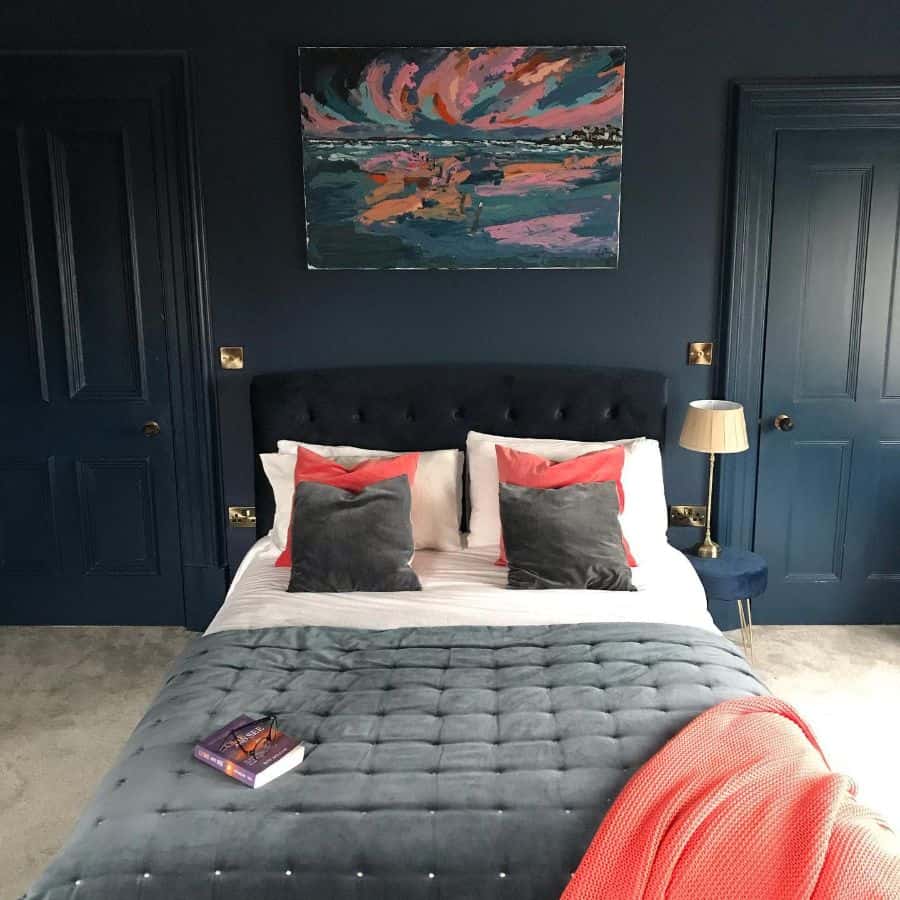 Dark blue bedroom with velvet bed, colorful artwork, and coral accents on pillows and throw.