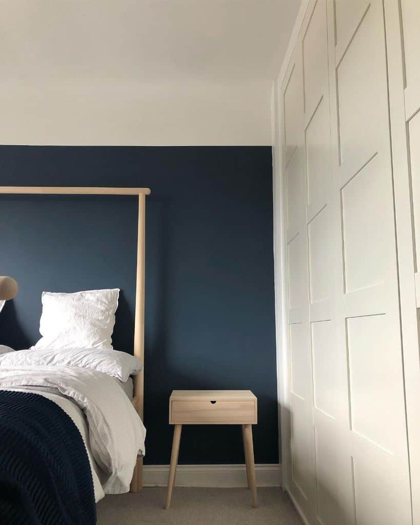 Minimalist bedroom with dark blue accent wall, wooden bed frame, and light wood nightstand.