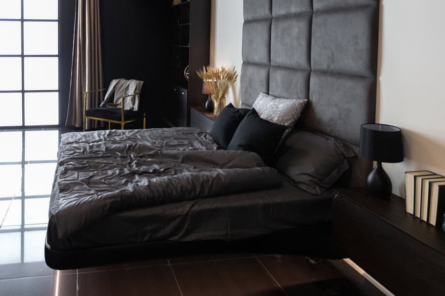 Modern bedroom with a dark color scheme, featuring a large bed with black bedding, a padded headboard, a nightstand with a lamp, and books on the shelf