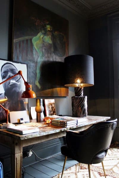 Eclectic workspace with a dark lamp, artwork, books, and a modern chair at a rustic desk