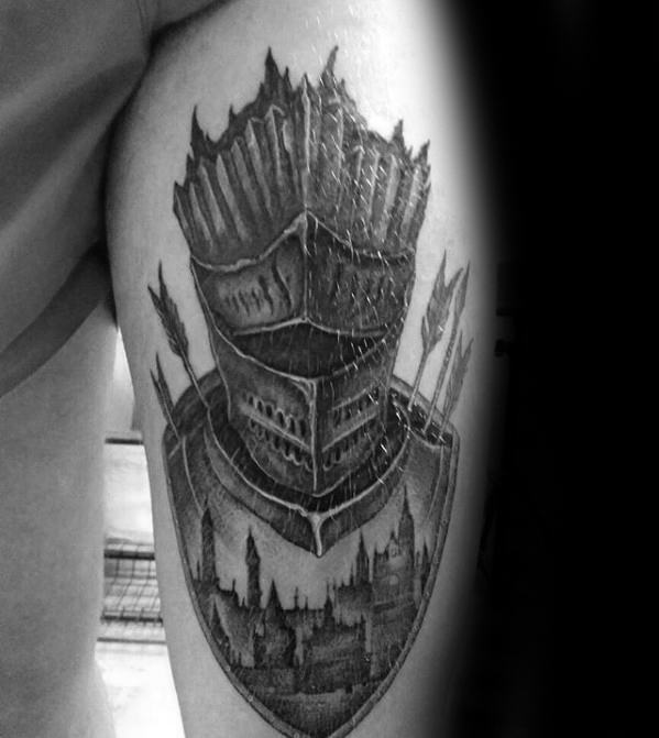 Black and grey Dark Souls tattoo of a knight’s helmet with arrows, set above a shield depicting a gothic cityscape.