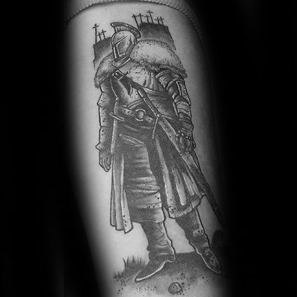 Detailed black and grey Dark Souls tattoo of a knight with a fur cloak, sword, and grave markers in the background.