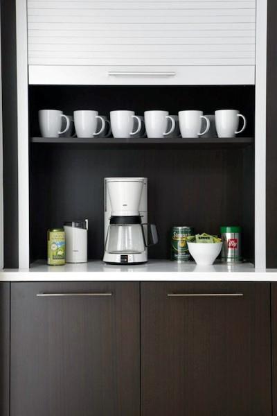 Compact coffee bar with dark cabinets, white mugs, and a drip coffee maker on open shelving.