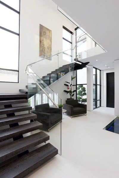Modern minimalist interior featuring a floating staircase, glass railing, and large windows with black couches and plants adding decor