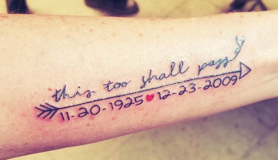 A "This Too Shall Pass" tattoo in cursive, with an arrow and heart, including dates "11-20-1925" and "12-23-2009" on a person's forearm.