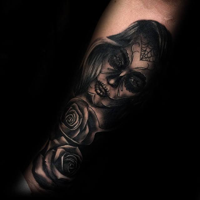 Day of the Dead-inspired tattoo featuring a woman's face with intricate sugar skull designs and black roses on the forearm