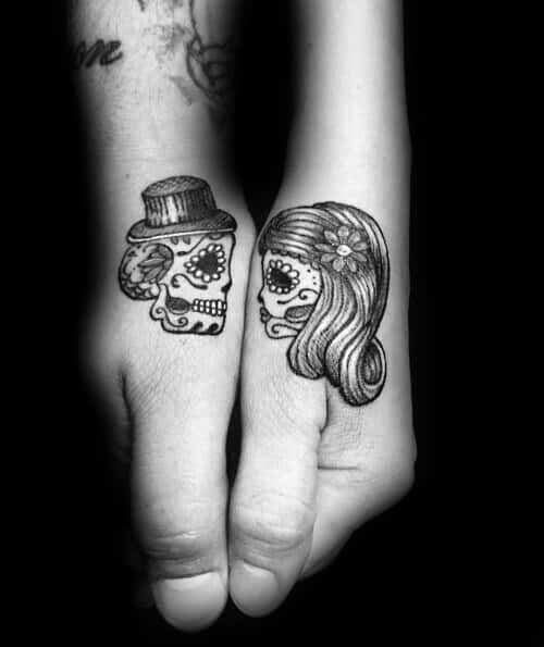 Matching sugar skull tattoos on thumbs, featuring a man with a hat and a woman with a flower, symbolizing eternal love and Día de los Muertos inspiration.