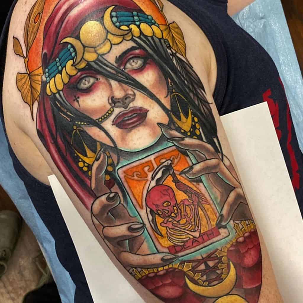 Bold and vibrant tattoo of a mystical gypsy woman with glowing eyes, intricate gold jewelry, and a 'Death' tarot card on the upper arm.