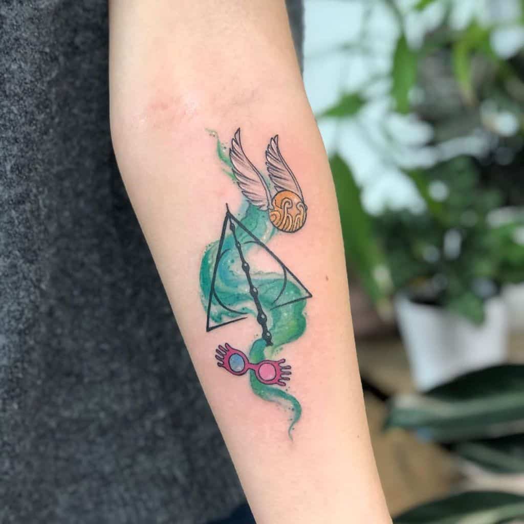 Colorful Deathly Hallows tattoo with a swirling green magical aura, Golden Snitch, and Luna Lovegood’s Spectrespecs on the forearm.