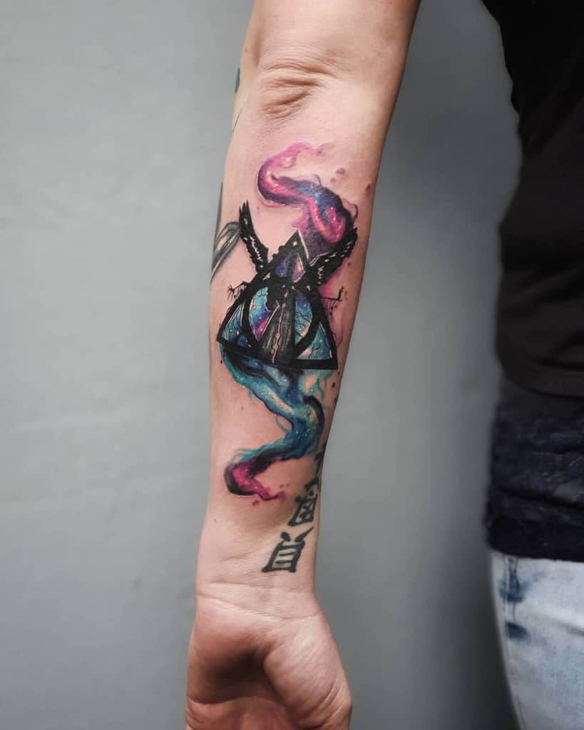 Vibrant Deathly Hallows tattoo with watercolor-style magical smoke in pink and blue, featuring a raven and Elder Wand details