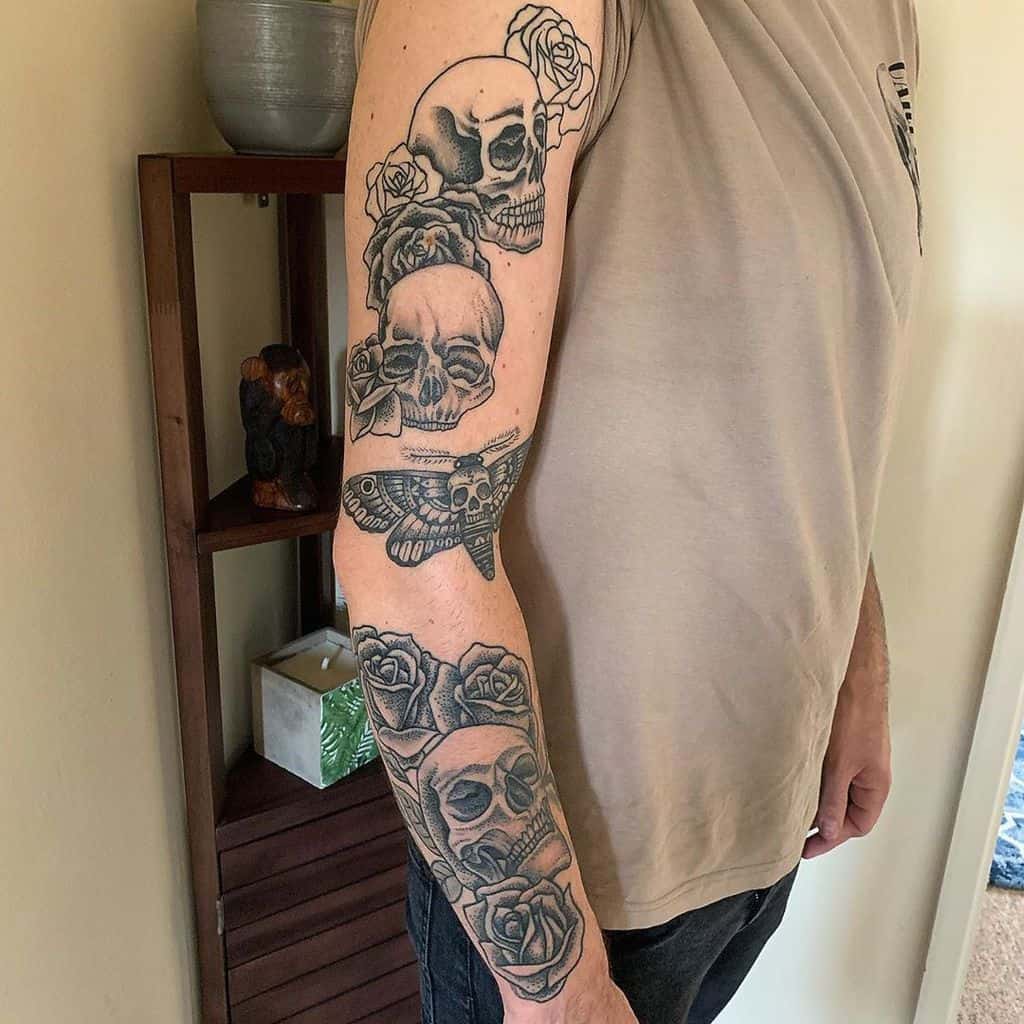 Black and grey tattoo sleeve featuring multiple skulls, roses, and a moth, creating a dark and symbolic design along the entire arm.