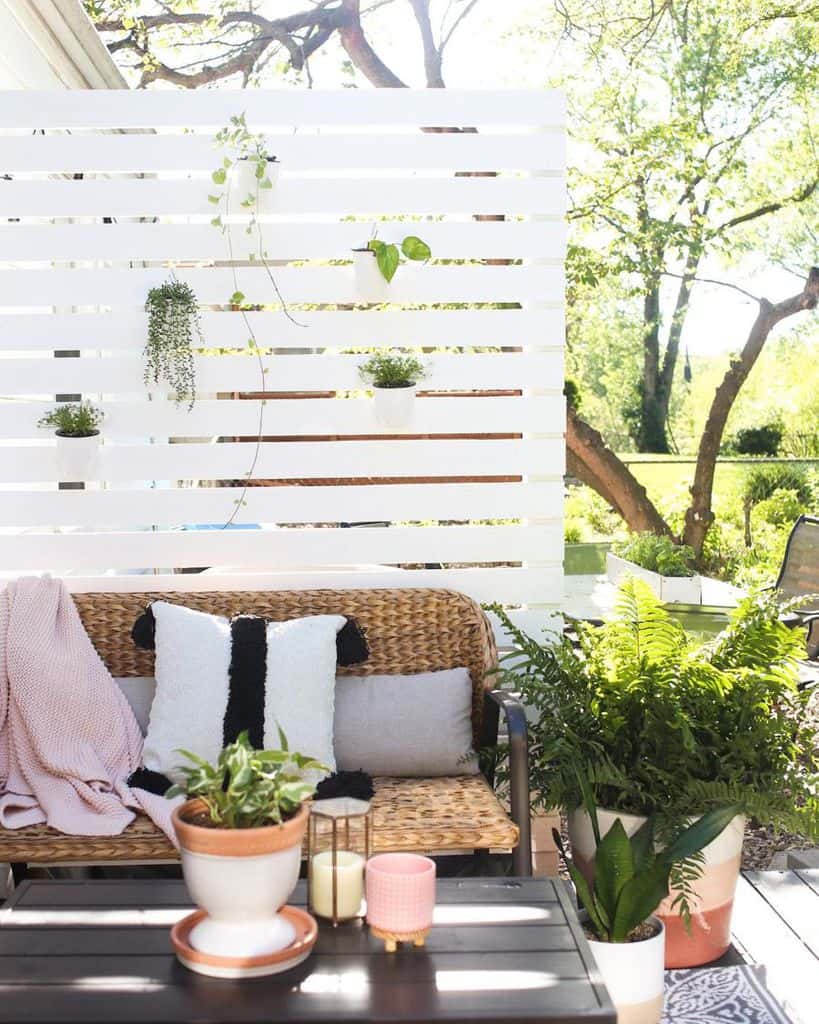 Bright outdoor patio with wicker sofa, white and gray pillows, plants on a wooden privacy screen, and a decorative coffee table
