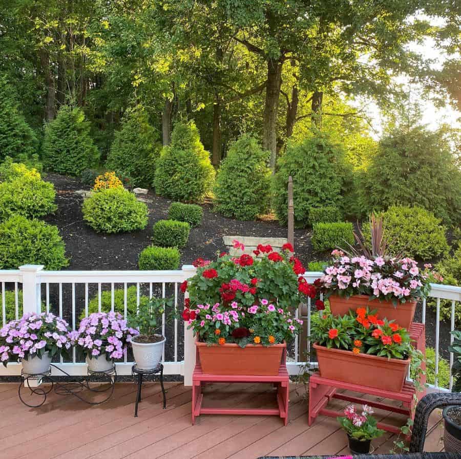 A sunny backyard deck with potted flowers in the foreground and lush green trees in the background