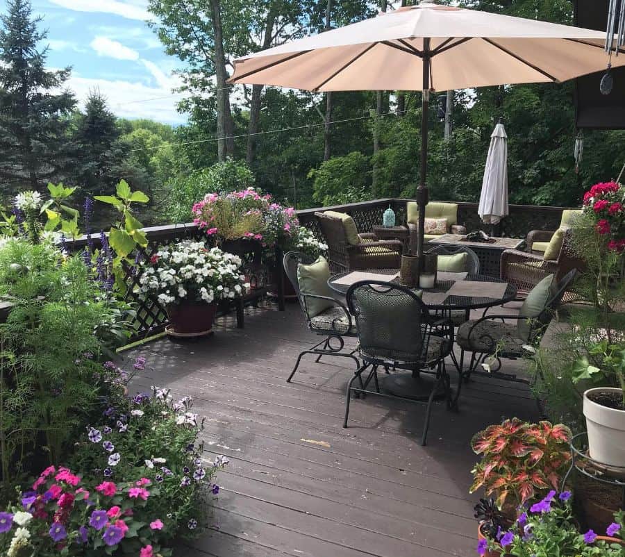 A wooden deck with patio furniture, an umbrella, and various potted flowers and plants in a lush, green garden setting