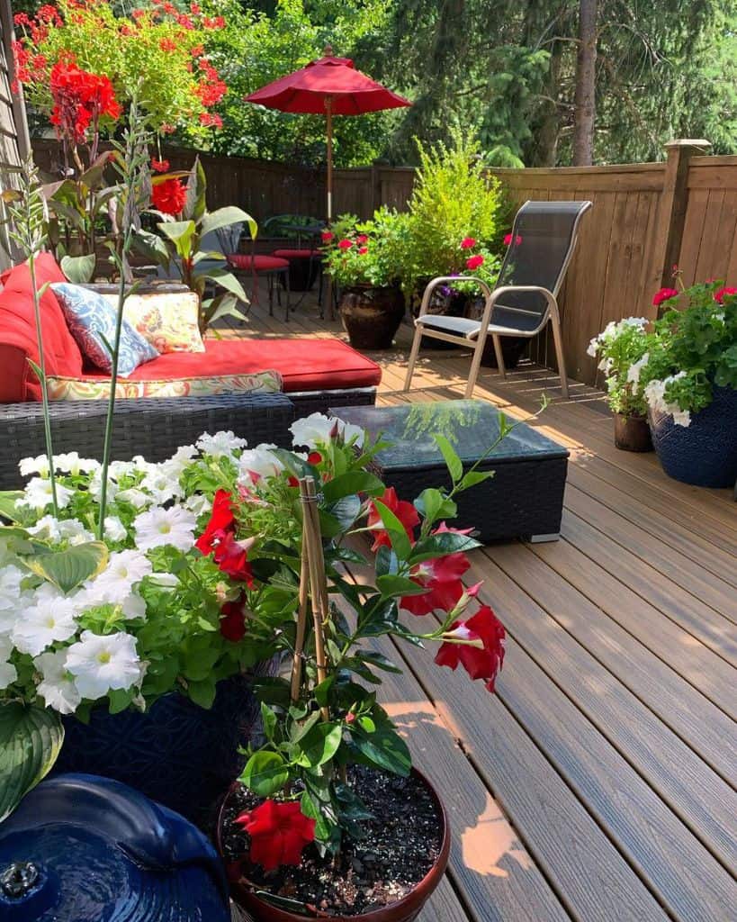 A cozy patio with red cushions, various potted plants, and a red umbrella, surrounded by a wooden fence and greenery