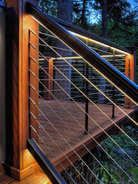 Elevated deck with cable railing, warm LED lighting, and forested background at dusk.