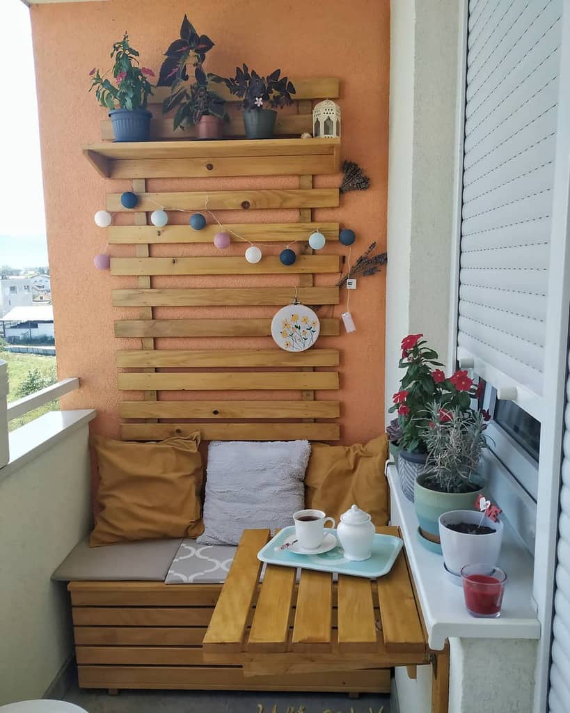 Small balcony with wooden seating, colorful cushions, and a decorative wall shelf with plants.