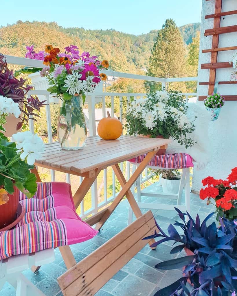 Colorful balcony with a wooden table, bright flowers, striped cushions, and a scenic mountain view.
