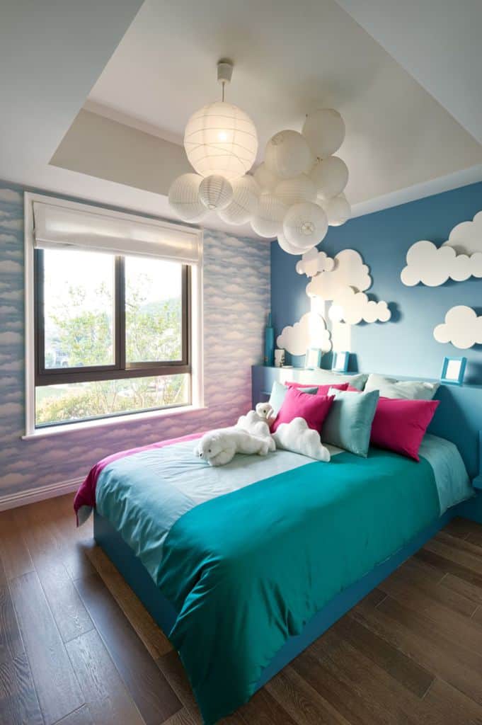 Whimsical bedroom with cloud-themed decor, teal bedding, pillows, and a plush toy on the bed under white hanging lights