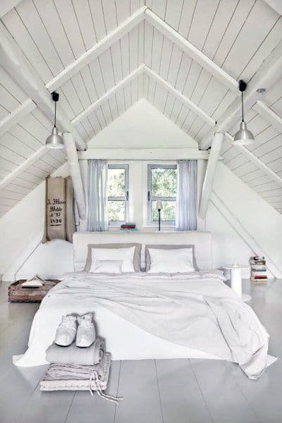 Cozy white attic bedroom with sloped ceiling, attic bed draped in white linens, and twin pendant lights