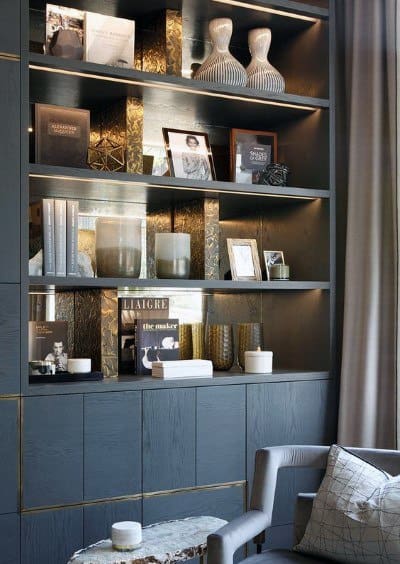 Modern living room with decorative shelves, books, vases, framed photos, and soft lighting above a gray armchair and table