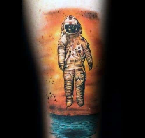 Deja Entendu astronaut tattoo showing a lone figure floating above the ocean with an orange sunset sky and scattered black birds.