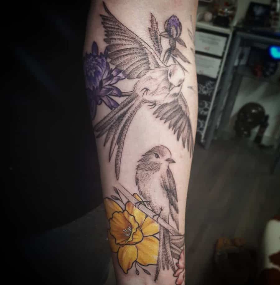 A detailed tattoo featuring two birds in flight, with flowers, including a yellow daffodil, elegantly incorporated into the design.
