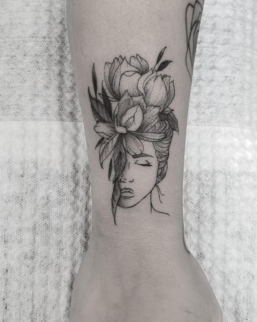 Elegant black and grey tattoo of a woman's face with closed eyes, adorned with detailed blooming flowers and leaves in her hair