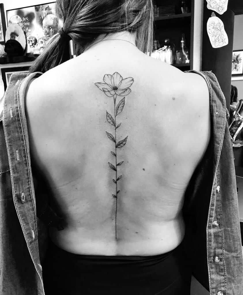 Minimalist fine-line tattoo of a single delicate flower with a long stem and leaves, running down the spine for an elegant look