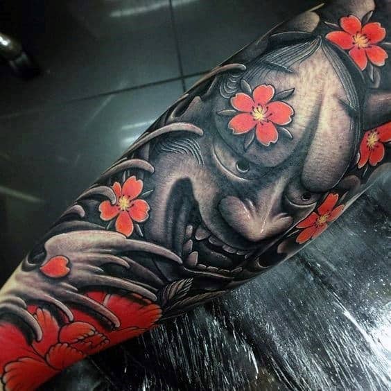 Realistic Japanese-style tattoo featuring a fierce Hannya mask with intricate shading, surrounded by vibrant red cherry blossoms