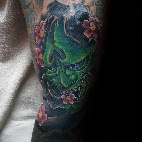 Japanese-style tattoo featuring a fierce green Hannya mask with intense shading, surrounded by pink cherry blossoms and dark swirling clouds